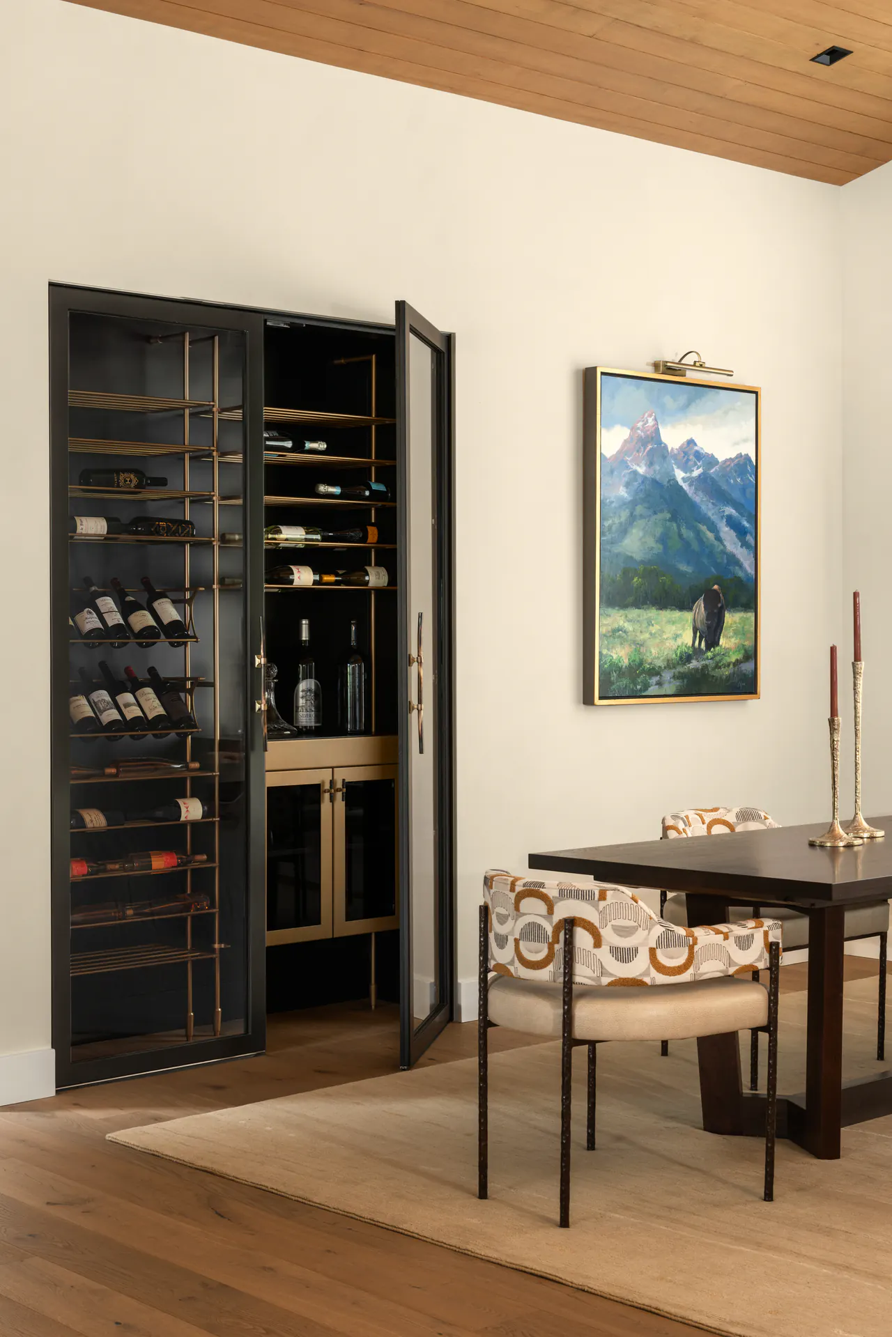 A dining room with a black steel wine cabinet built into a pale wall, one glass door open, beside a table and a landscape painting.