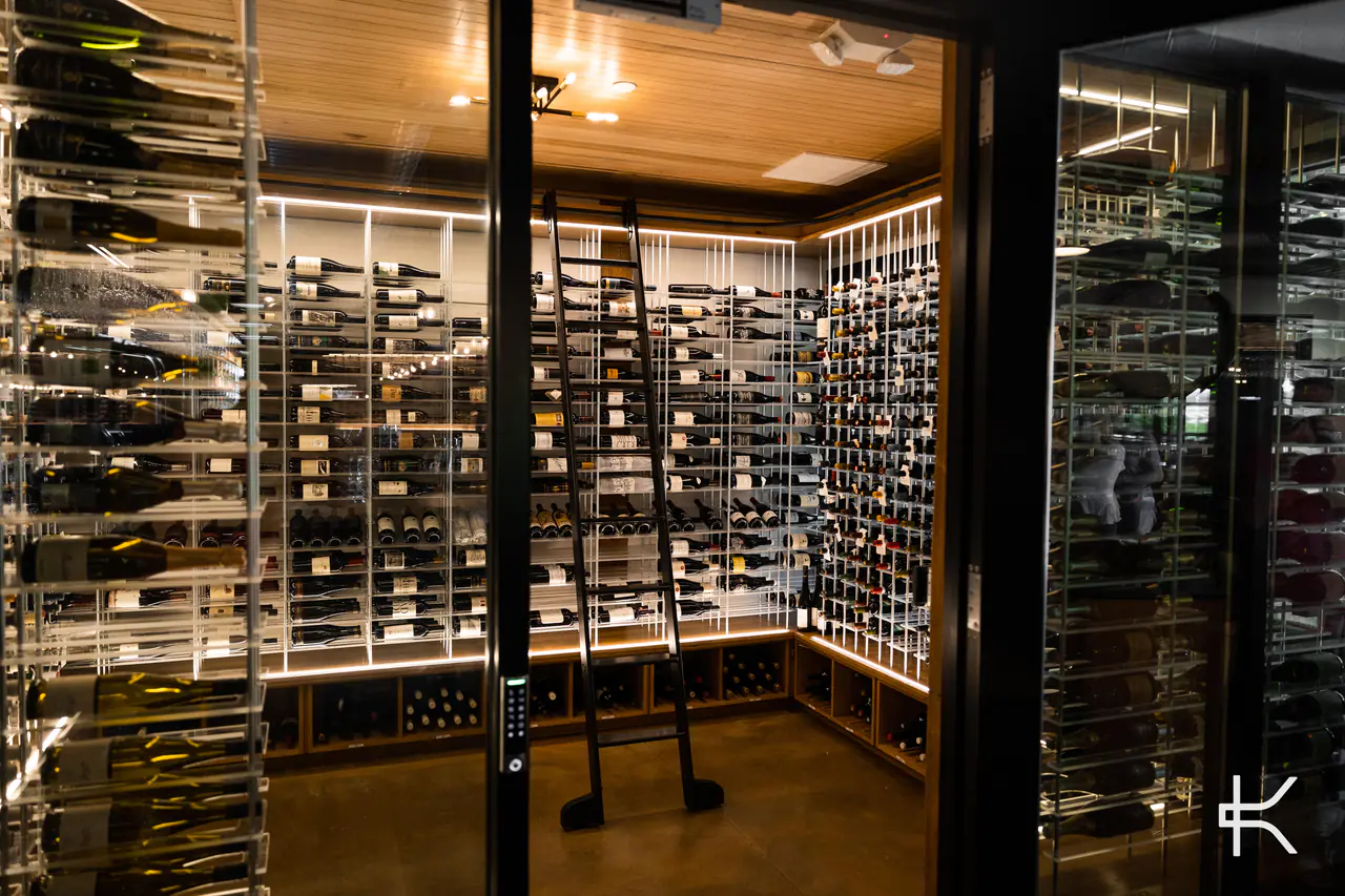 Looking through a glass wall into a wine room lined with lit white racks and a library ladder.