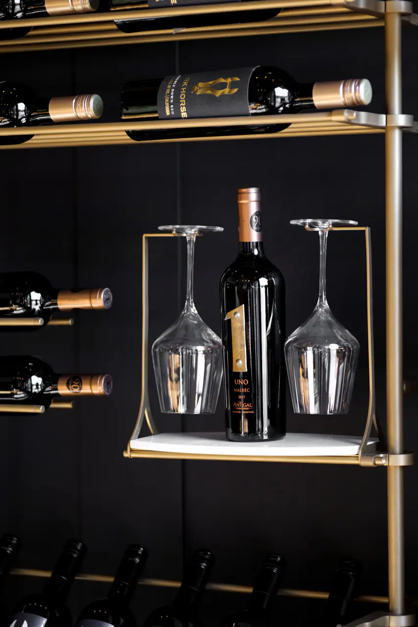 A bottle standing between two hanging glasses on a brass-framed shelf.