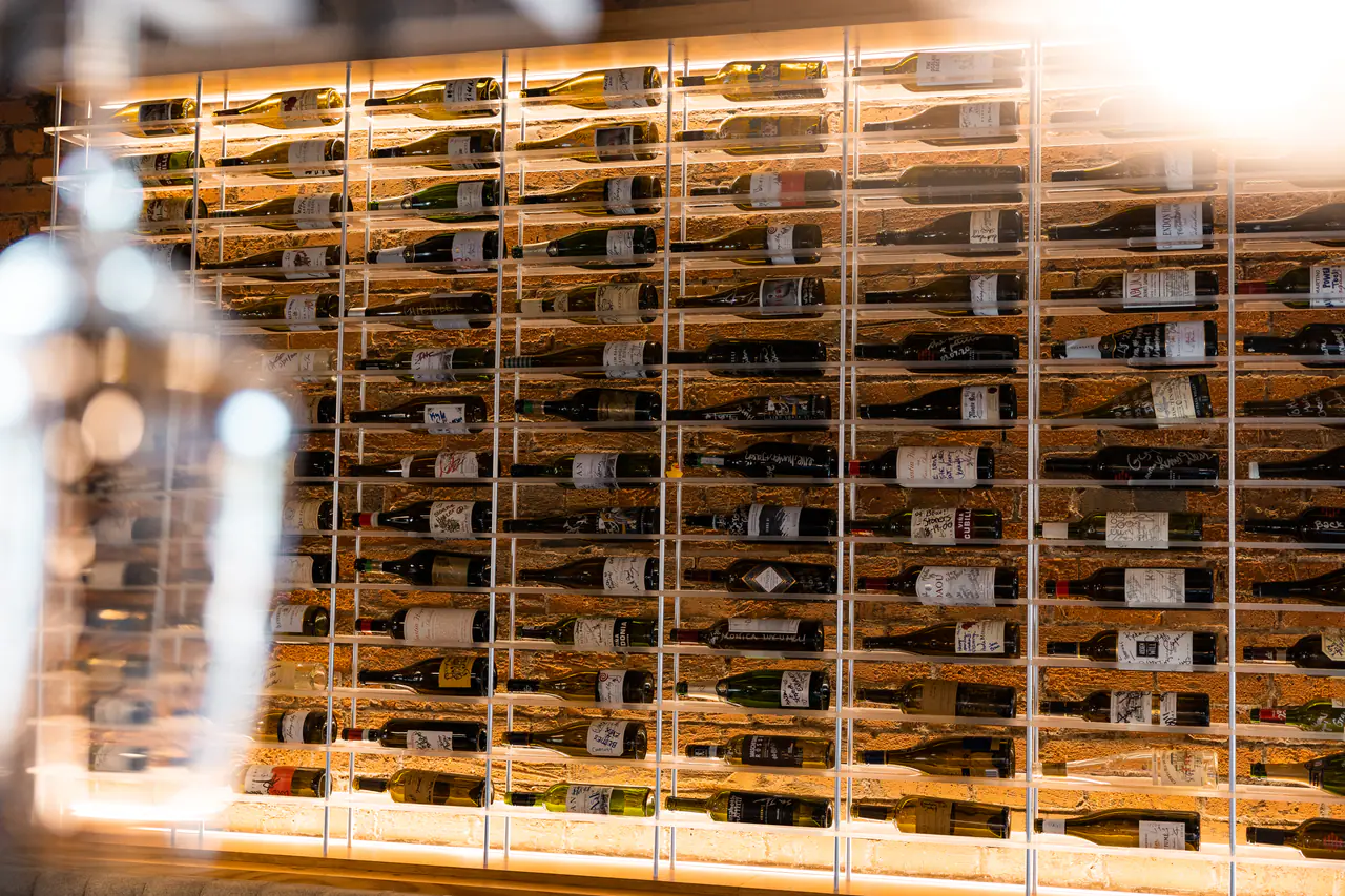 A wall of wine in a white steel grid, lit along its base, seen past a glass.