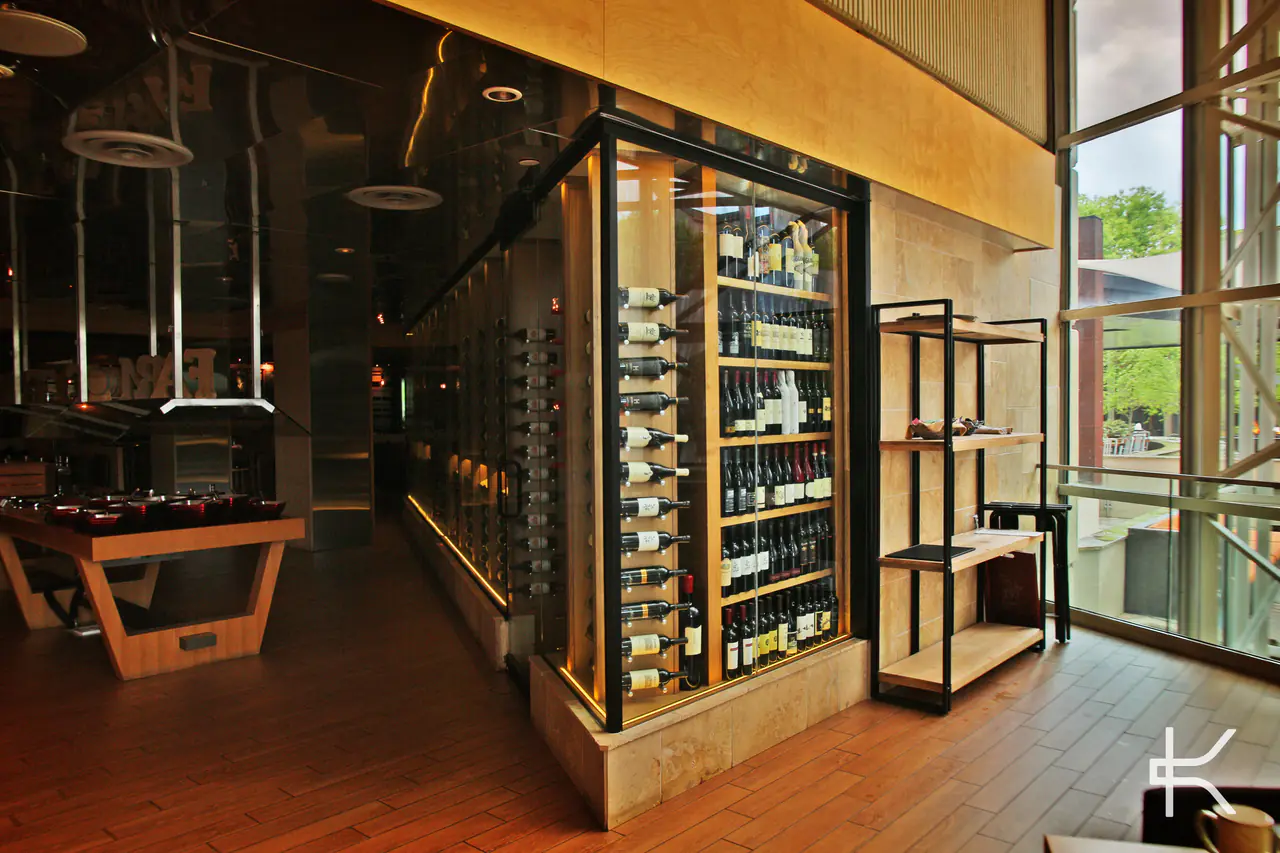 A glass-walled wine room with timber shelving beside a tall window, a long table in the room beyond.