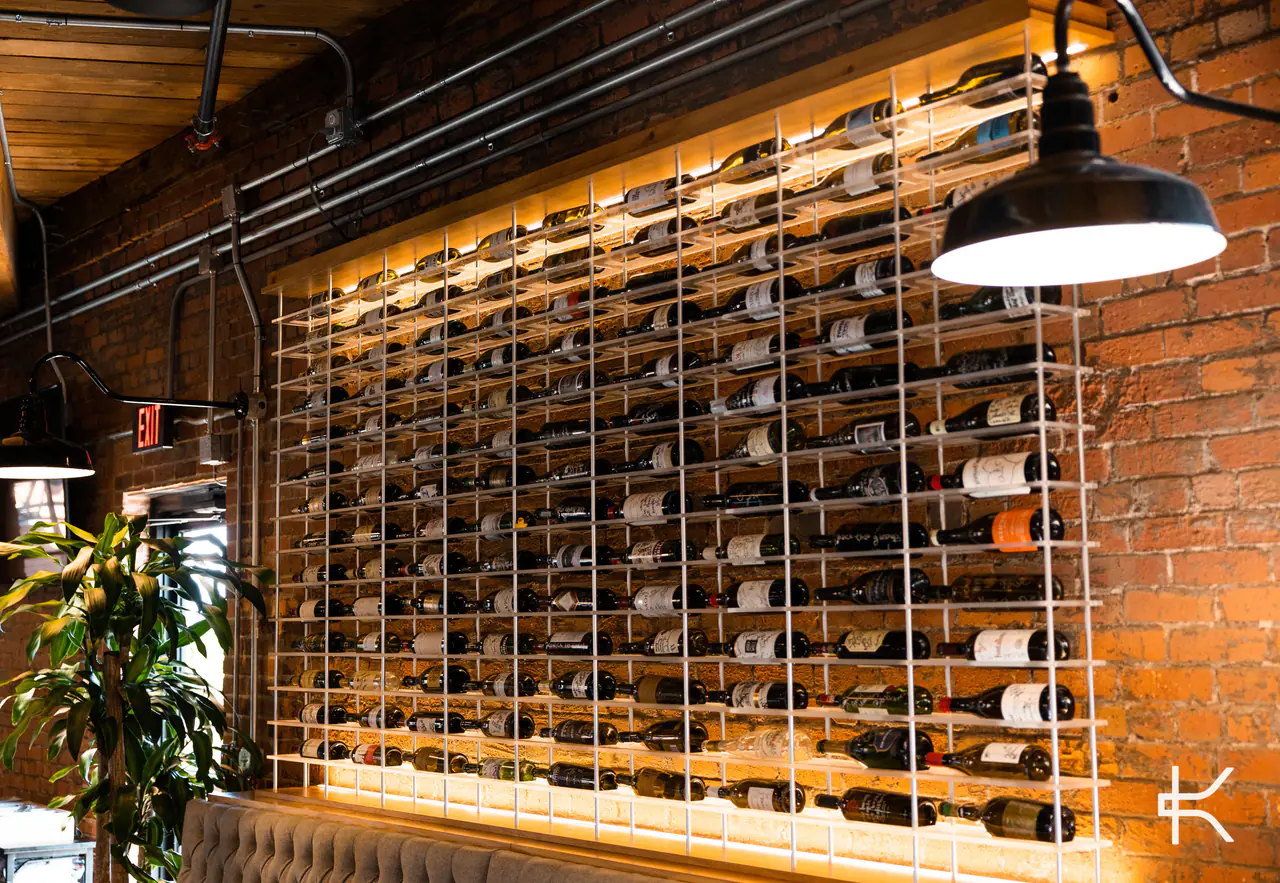 A white steel wine grid against a brick wall, lit from behind, beneath an enamel pendant lamp.
