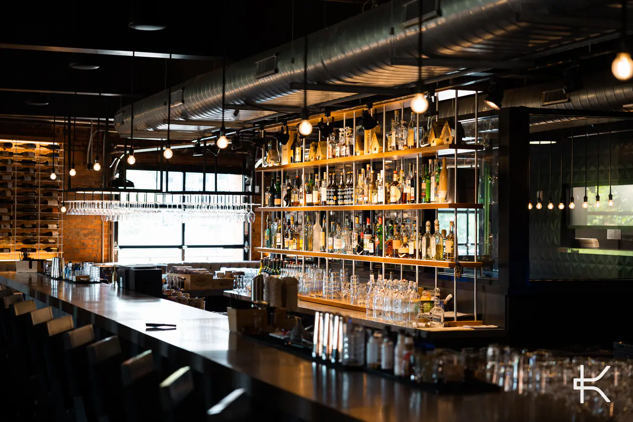 A long bar with an open back bar of spirits and hanging glassware, windows beyond.