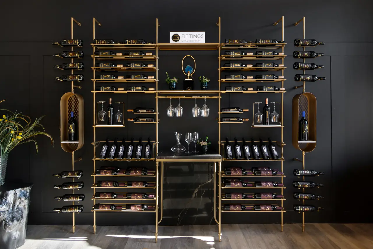 A full wine wall of brass uprights, bottles on pegs, glass shelves, two capsule niches and a cabinet.