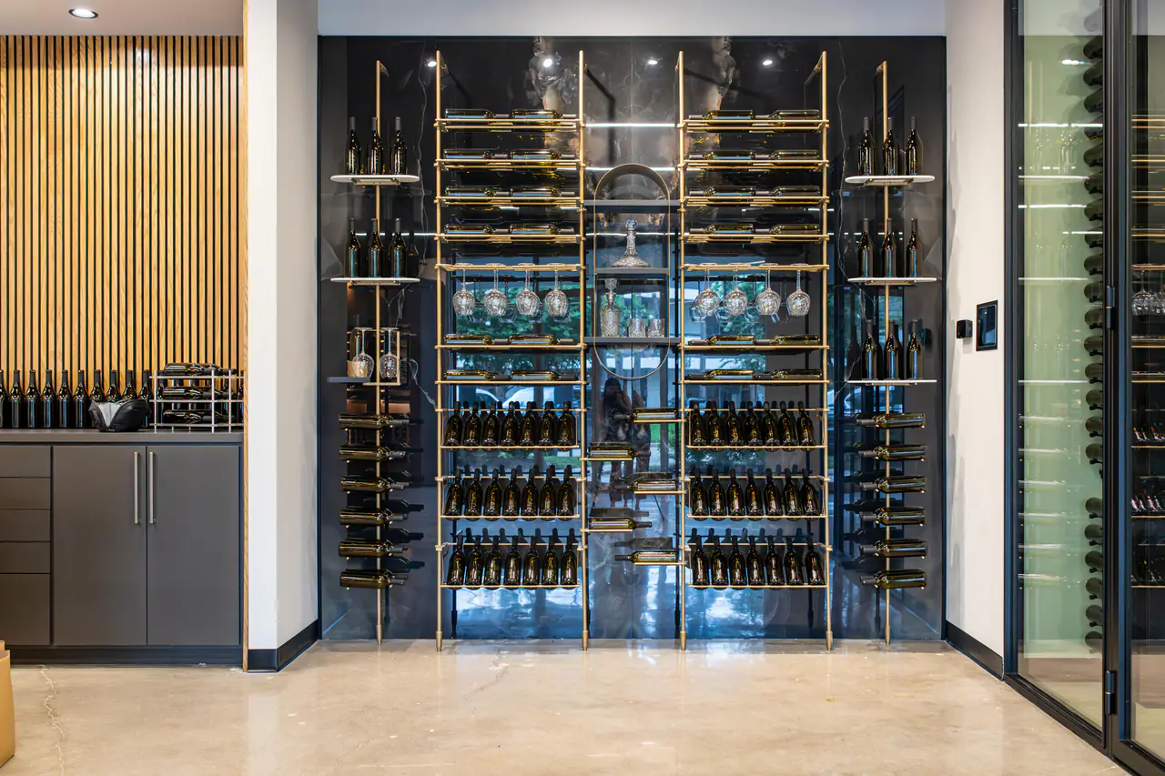A brass wine wall of racks, glass shelves and hanging glasses against black stone, a glass wine room at right.