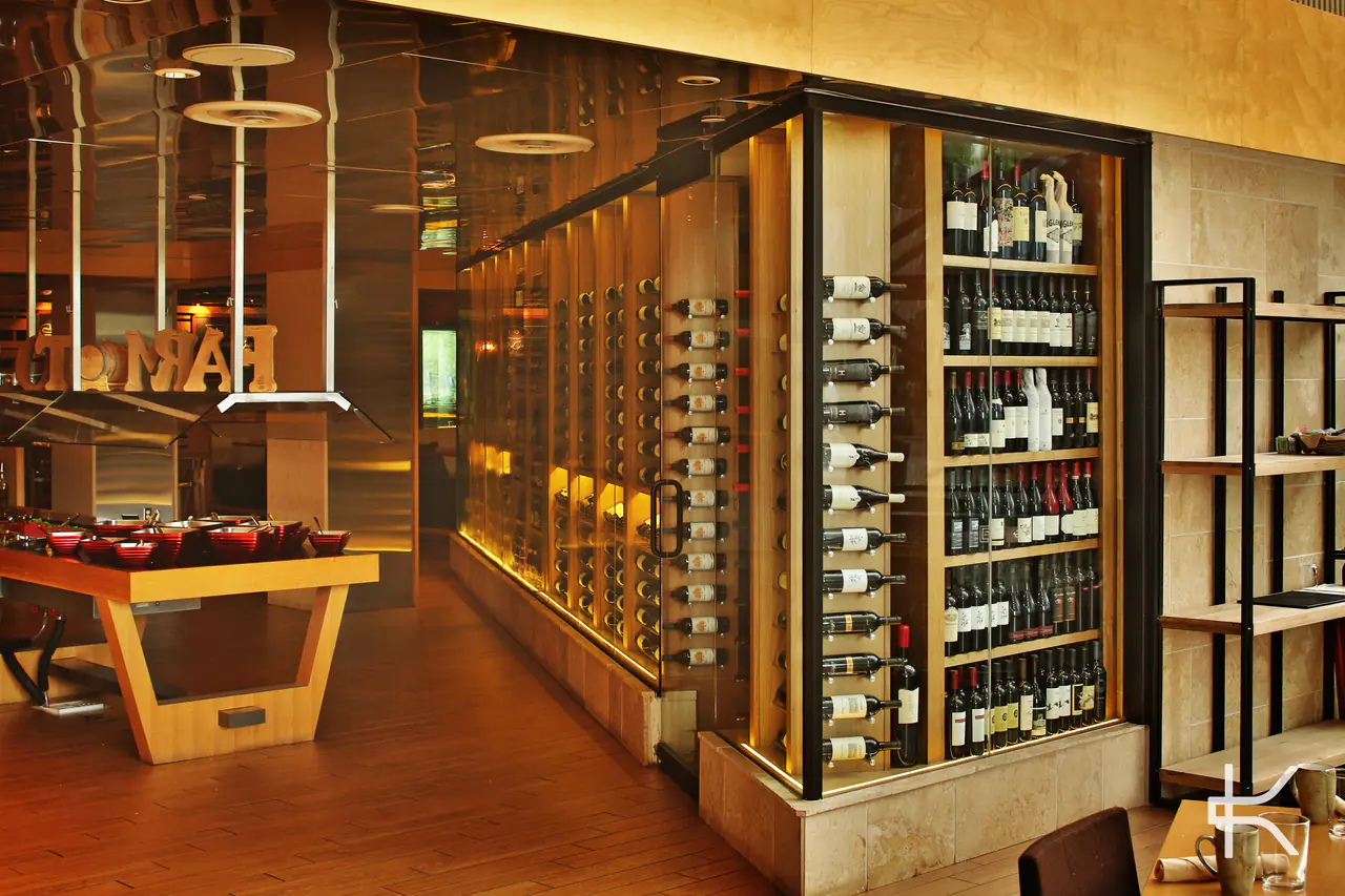 The corner of a glass-walled wine room, timber racks lit behind black steel frames.