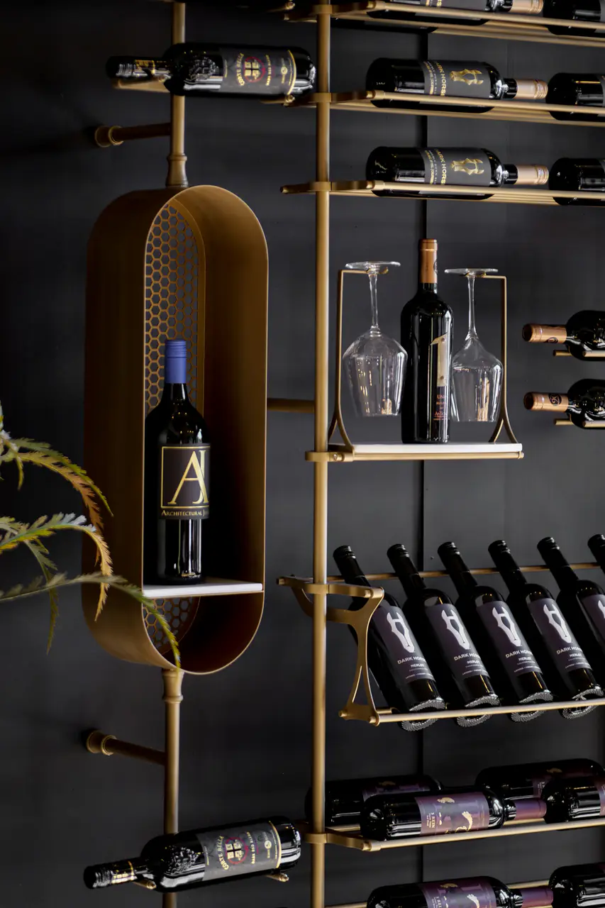 A brass wine wall with a capsule niche, a glass shelf and bottles on pegs.