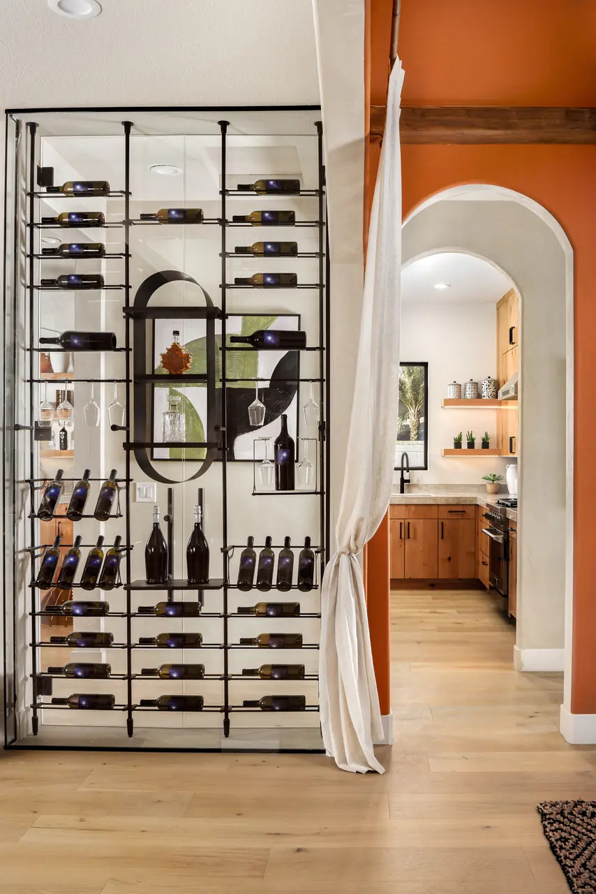 A black steel wine wall with an oval capsule shelf, beside an arched doorway into a terracotta kitchen.