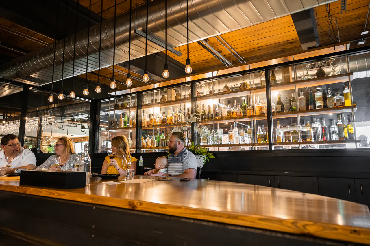 Guests at a long timber bar before a glass-fronted back bar of lit spirits.