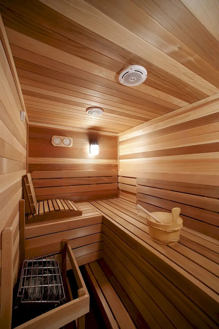 A cedar-lined sauna with slatted benches, a wall lamp and a thermometer by the door.